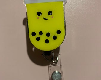 May include: A yellow boba tea charm with a smiling face and black boba pearls. The charm is attached to a clear plastic retractable badge holder with a silver clip and a glittery white button.