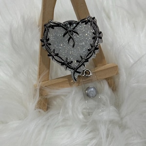 May include: A heart-shaped badge reel with a glittery silver center and a black barbed wire design around the edges. The reel has a clear plastic retractable cord and a silver clip. The badge is displayed on a small wooden easel.