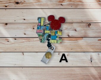 May include: A colorful 'LOVE' charm with a red Mickey Mouse head on top, attached to a silver retractable badge holder with a gold accent. The charm is made of a glittery material and features a game board design.