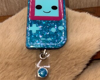 May include: A blue glittery badge reel with a retro video game design. The game console has a light blue screen with a smiling face and a pink border. The reel has a clear plastic strap and a silver clip.