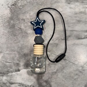 May include: An air freshener with a clear glass bottle and a black cord. The top features a navy blue star, blue and gray beads. The cord has a black magnetic clasp.
