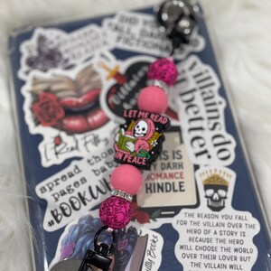 May include: A pink and black phone charm with a skull and crossbones design and the text "Let Me Read In Peace". The charm is made of beads and has a metal clip. The charm is on a blue background with other stickers.