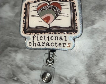 May include: A white and silver badge holder with a book and heart design. The book has a brown and black leopard print cover and the heart is filled with different colored patterns. The text on the badge holder reads "in love with fictional characters".