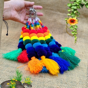 May include: A collection of keychains featuring multiple rows of vibrant, rainbow-colored pom-poms arranged in vertical strands. Each strand is topped with a metal ring and finished at the bottom with a long, colorful thread tassel.