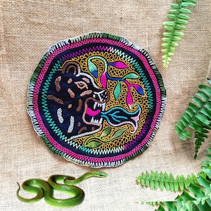 Peut inclure: Un écusson brodé circulaire représentant une tête de panthère noire avec la gueule ouverte et une flamme stylisée, entourée de motifs floraux et de feuilles colorés en rose, jaune et vert, avec une bordure cousue blanche sur fond sombre.