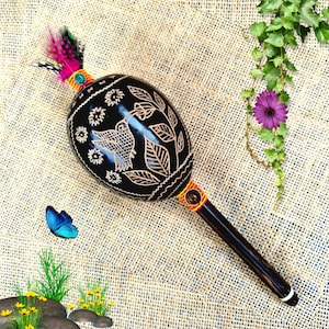 Puede incluir: Una maraca negra con un diseño grabado de un colibrí y flores en color beige claro. El mango y la parte superior están decorados con bandas tejidas en naranja y amarillo, una pequeña piedra verde y un grupo de plumas rosas, negras y blancas.