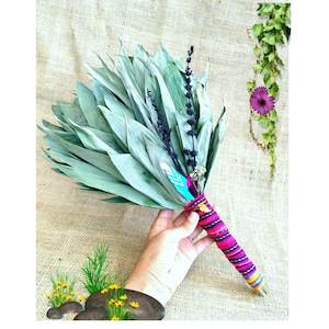 Può includere: Un mazzo di foglie verdi essiccate e steli di lavanda viola legato con un tessuto intrecciato dai colori vivaci rosa, nero e giallo. Un piccolo dettaglio con una piuma turchese e bianca è inserito nella legatura della composizione.