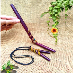 Puede incluir: Una varita de madera morada larga y delgada con una escultura de serpiente marrón enroscada y un pequeño acento de piedra azul. Debajo, un colgante de madera morada a juego cuelga de un cordón negro, decorado con una serpiente amarilla y un detalle floral.