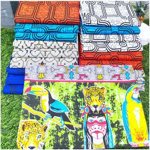 Puede incluir: Montones de tela colorida con varios patrones. Los diseños incluyen formas geométricas en naranja, azul y blanco, y una impresión vibrante con un tucán, un jaguar y una mujer indígena. Las telas están cuidadosamente dobladas, mostrando una variedad de colores y diseños.