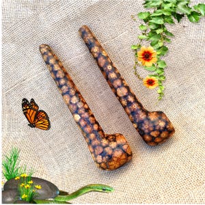 Pode incluir: Duas piteiras de madeira com um padrão texturizado único em tons de castanho e bege. As piteiras têm uma forma curva com um caule e uma tigela. O fundo apresenta uma borboleta, flores e uma cobra.