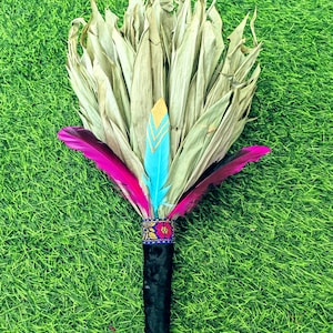 Puede incluir: Un palo de limpieza de hierbas secas y plumas con un mango de terciopelo negro y un adorno floral colorido. Las plumas son rosas, azules y doradas.