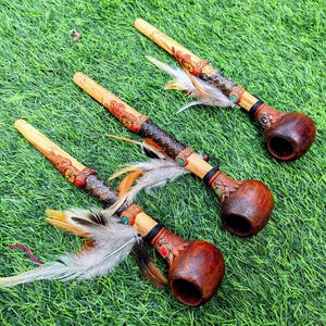 Puede incluir: Tres pipas de madera hechas a mano con tallas decorativas y adornos de plumas descansan sobre hierba verde. Las pipas tienen un cuenco marrón oscuro, un tallo de color claro y están adornadas con plumas de colores.
