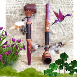Pode incluir: Duas cachimbos de madeira artesanais com detalhes decorativos. Um cachimbo tem uma tigela arredondada e o outro um design reto. Ambos os cachimbos apresentam detalhes em couro, adornos de penas e enfeites de pedras preciosas. Um beija-flor paira perto.