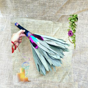 May include: A bundle of dried sage leaves with a black fabric-wrapped handle, decorated with bright pink and turquoise feathers and a patterned fabric accent. The bundle is positioned near a set of three cream-coloured pillar candles.
