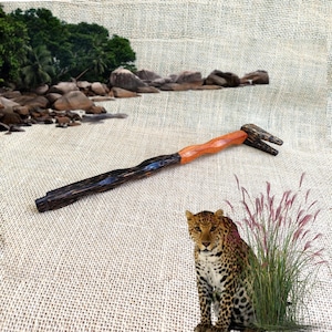 May include: A hand-carved wooden tool with a dark, textured handle and a lighter, orange-toned central section. The forked end is also dark. The background features a leopard, tall grass, and a beach scene.