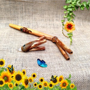 Puede incluir: Una flauta de madera con acabado marrón y beige, con boquilla decorativa y sección de tres agujeros. La flauta va acompañada de una pieza de madera separada, tallada intrincadamente. Una mariposa azul descansa cerca, con girasoles en primer plano.