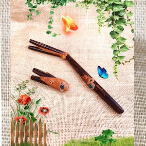 Op de afbeelding: Twee houten haarstokjes met decoratieve accenten. Eén stokje heeft een slangenontwerp met turkooizen stenen, de andere een oogvormige versiering. De stokjes zijn donkerbruin met een natuurlijke houtnerf.