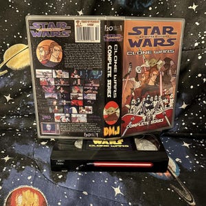 May include: A Star Wars: Clone Wars Complete Series DVD set and a VHS tape. The DVD case features animated characters and the text "Star Wars Animated Classics." The VHS tape is black with red accents and the text "Star Wars Clone Wars."
