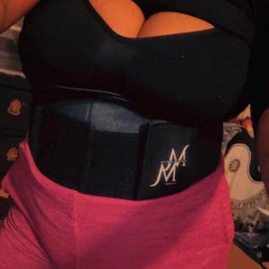 Könnte beinhalten: Ein schwarzer Taillentrainer mit den weißen kursiven Buchstaben "M" auf der Vorderseite. Der Taillentrainer wird über einem schwarzen Oberteil und einer rosa Hose getragen. Der Taillentrainer scheint aus einem dicken, strapazierfähigen Material zu bestehen.