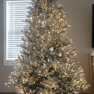 May include: A flocked artificial Christmas tree with white lights and a wooden tree skirt that says "Merry Christmas".