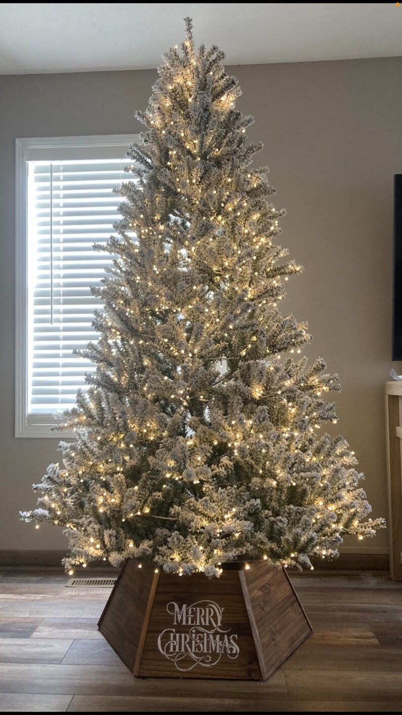 May include: A flocked artificial Christmas tree with white lights and a wooden tree skirt that says "Merry Christmas".