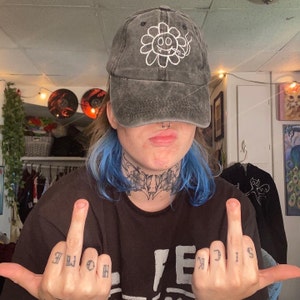 May include: A person with blue hair is wearing a black baseball cap with a white flower design. The cap has the text "OO" inside the flower. The person is wearing a black shirt with a white lightning bolt design and the text "FE" and "IG" on their fingers.