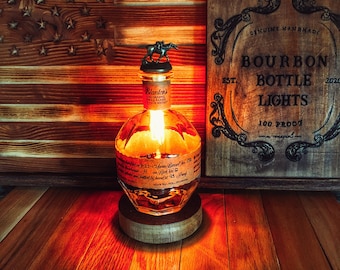 May include: A repurposed bourbon bottle lamp with a wooden base and a horse-shaped bottle stopper. The bottle is clear glass with a label that reads "Blanter's The Original Single Barrel Bourbon Whiskey". The lamp is lit and casting a warm glow. The background is a wooden surface with an American flag and a sign that reads "Genuine Handmade Bourbon Bottle Lights Est. 2020 100 Proof".