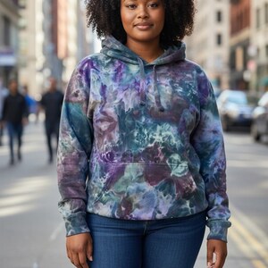May include: A tie-dye hoodie in shades of purple, blue, and green. The hooded sweatshirt has a front pocket and long sleeves. The person in the image is wearing blue jeans. The background is a city street.