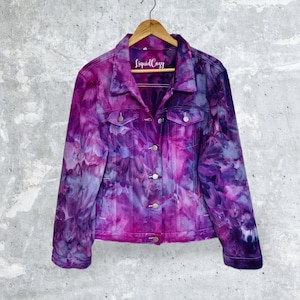 K&ouml;nnte beinhalten: Lilafarbene Batik-Jeansjacke mit einem Holzb&uuml;gel und einem Etikett mit der Aufschrift "Liquid Cozy".