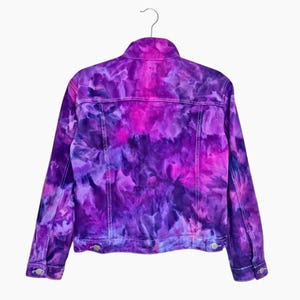 K&ouml;nnte beinhalten: Eine lilafarbene Batik-Jeansjacke im klassischen Jeansjackenstil. Die Jacke hat ein helllila, dunkellila und rosafarbenes Batik-Muster.