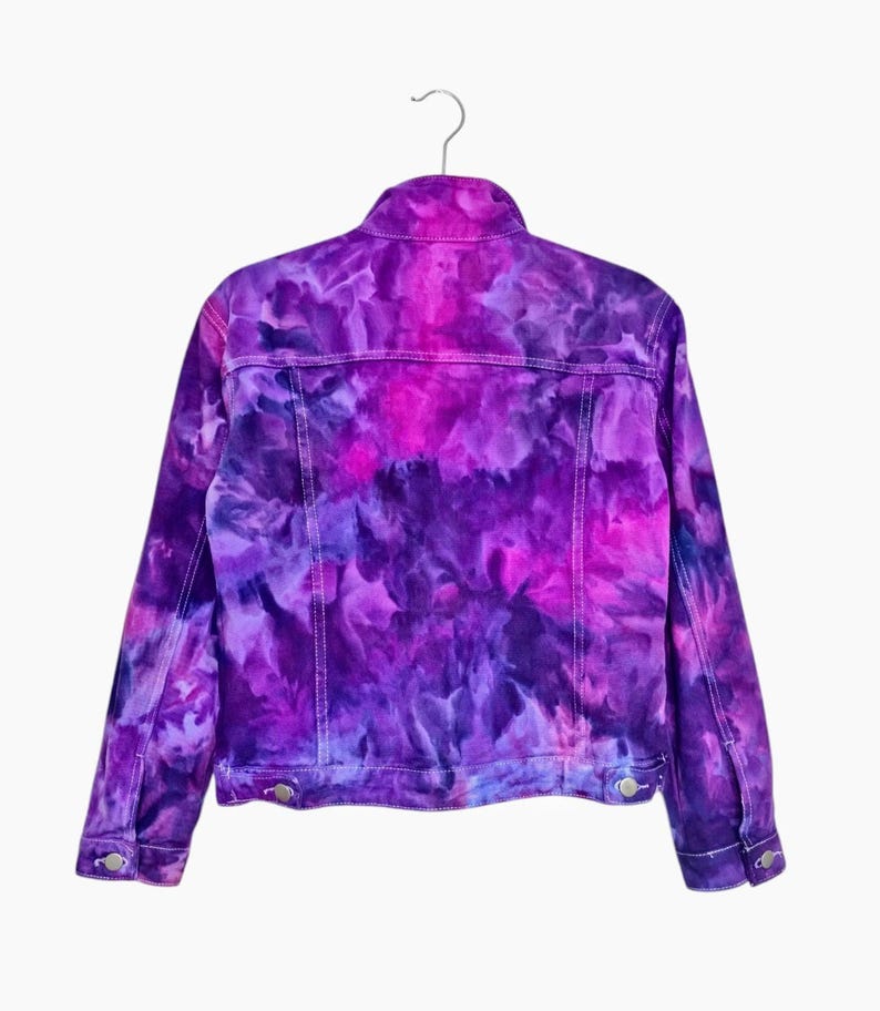 K&ouml;nnte beinhalten: Eine lilafarbene Batik-Jeansjacke im klassischen Jeansjackenstil. Die Jacke hat ein helllila, dunkellila und rosafarbenes Batik-Muster.