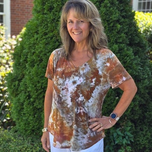 May include: A tie-dye V-neck t-shirt in shades of brown, white, and rust. The short-sleeved shirt has a relaxed fit and is paired with white pants. The model is wearing a watch and bracelet.