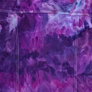 K&ouml;nnte beinhalten: Lilafarbene Tie-Dye-Jeansjacke mit wei&szlig;en N&auml;hten. Die Jacke hat ein verblichenes, aquarellartiges Design.