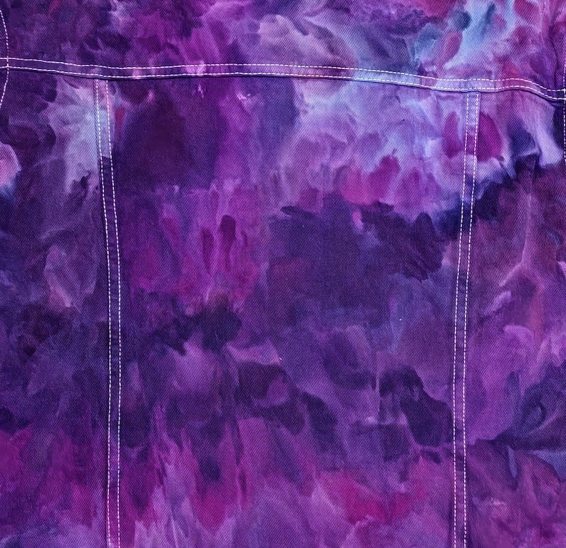 K&ouml;nnte beinhalten: Lilafarbene Tie-Dye-Jeansjacke mit wei&szlig;en N&auml;hten. Die Jacke hat ein verblichenes, aquarellartiges Design.