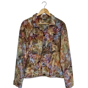 Könnte beinhalten: Eine langärmelige Jeansjacke mit einem mehrfarbigen Batikmuster in Braun-, Lila-, Blau- und Grüntönen. Die Jacke verfügt über einen klassischen Kragen, eine Knopfleiste vorne, zwei Brusttaschen mit Knopfklappen und geknöpfte Ärmelbündchen.