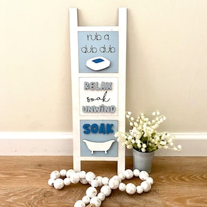 May include: White wooden ladder with three blue and gray signs. The top sign says "rub a dub dub" with a blue bathtub icon. The middle sign says "relax soak unwind". The bottom sign says "soak" with a blue bathtub icon.