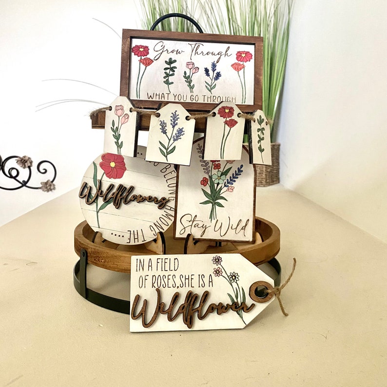 May include: A set of wooden signs with floral designs and inspirational quotes. The signs feature phrases like "Grow Through What You Go Through", "Wildflowers Belong Among The", "Stay Wild", and "In a Field of Roses, She is a Wildflower".