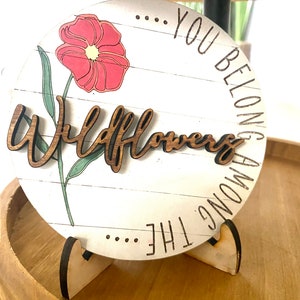 May include: A round wooden sign with a white background and a pink flower graphic. The sign reads "... You Belong Among The Wildflowers".