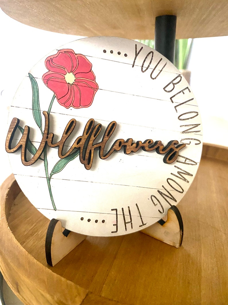 May include: A round wooden sign with a white background and a pink flower graphic. The sign reads "... You Belong Among The Wildflowers".