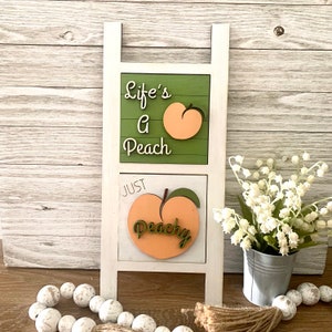 May include: White wooden ladder with two shelves. The top shelf has a green background with the text "Life's a Peach" and a peach graphic. The bottom shelf has a white background with the text "Just Peachy" and a peach graphic.