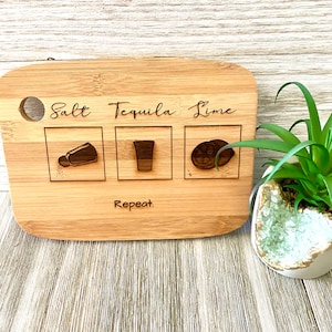 May include: A wooden cutting board with the words "Salt Tequila Lime" and "Repeat" engraved on it. The board has three sections with illustrations of a salt shaker, a shot glass, and a lime wedge.