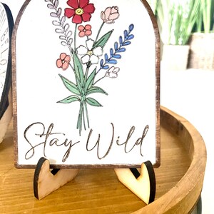 May include: A white wooden sign with a floral design and the text "Stay Wild". The sign is on a wooden stand.