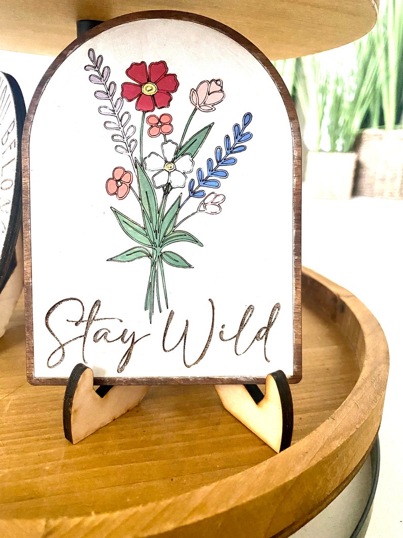 May include: A white wooden sign with a floral design and the text "Stay Wild". The sign is on a wooden stand.