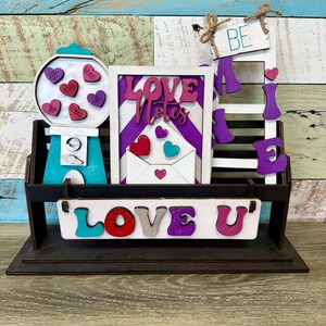 May include: A wooden Valentine's Day decoration with a gumball machine, a sign saying "Love Notes", and a bench with "Be Mine" and "Love U". The decoration is painted in teal, purple, and pink. A charming gift for a loved one.