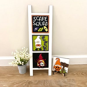 May include: White wooden ladder shelf with three Halloween themed signs. The top sign reads "Scare Squad" in black letters with orange and black candy and a spider. The middle sign has a green background with a white gnome holding a bag of candy and the letters "EEK" in purple. The bottom sign has a black background with a red gnome holding a pitchfork and a skull. The bottom sign also has a small wooden sign that reads "Trick or Treat".