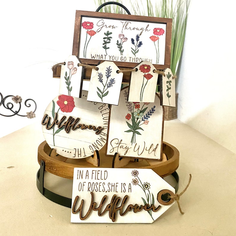 May include: A set of wooden signs with floral designs and inspirational quotes. The signs feature phrases like "Grow Through What You Go Through", "Stay Wild", and "In a Field of Roses, She is a Wildflower".