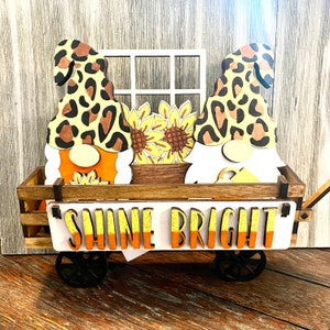 May include: A wooden wagon with a white sign that reads "Shine Bright". The wagon is decorated with two gnomes wearing leopard print hats and holding sunflowers.