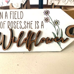 May include: A white wooden sign with the text "In a field of roses, she is a Wildflower" in brown lettering. The sign has a brown wooden circle attached to a string and a floral design with pink and yellow flowers.