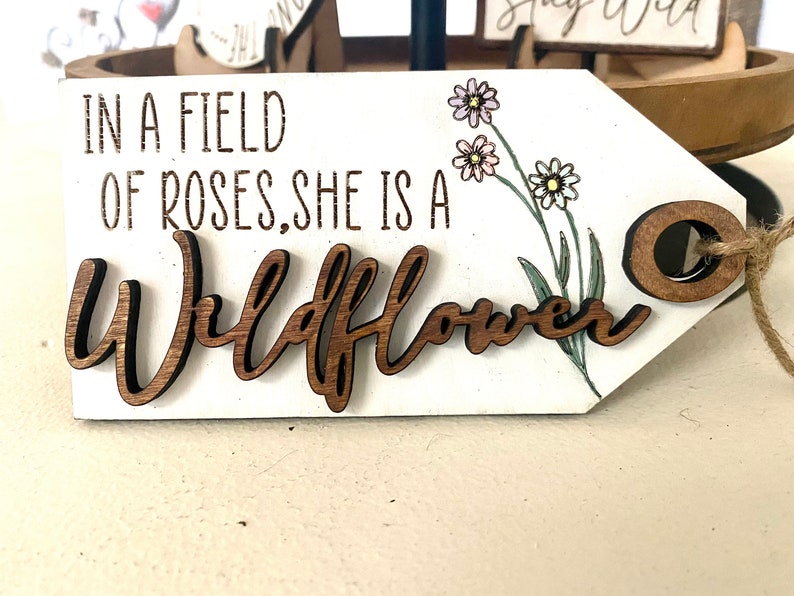 May include: A white wooden sign with the text "In a field of roses, she is a Wildflower" in brown lettering. The sign has a brown wooden circle attached to a string and a floral design with pink and yellow flowers.