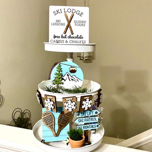 May include: A white tiered tray decorated for a ski lodge theme. The top tier has a sign that reads "SKI LODGE" with crossed skis and the text "SKI LESSONS", "GUIDED TOURS", "free hot chocolate", and "CABINS & CHALETS". The middle tier has a blue and white round sign with a mountain scene and a ski lift. The bottom tier has a blue and white sign with the text "SKI LIFT", "SKI PATROL", and "SKI RENTALS".